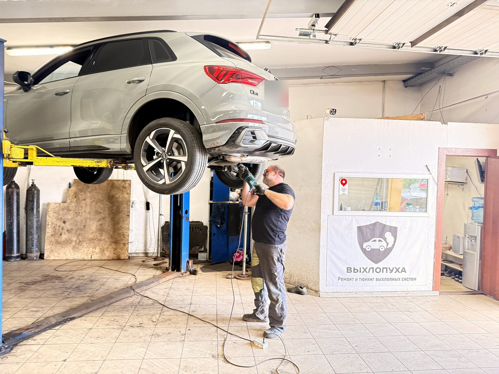 Серая Audi Q3 на подъёмнике в цехе «Выхлопухи» на Южном шоссе, мастер под машиной снимает штатную банку, рядом баннер «Выхлопуха»