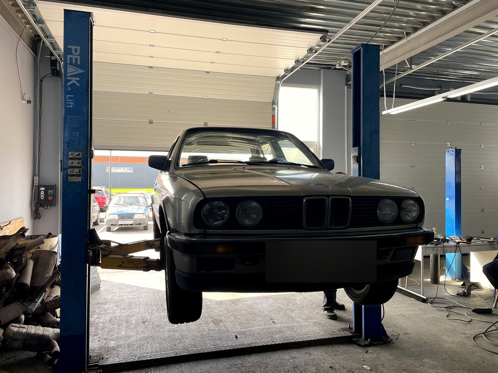 Серебристая BMW 325e в кузове E30 поднята на подъёмнике в боксе «Выхлопухи», вид спереди