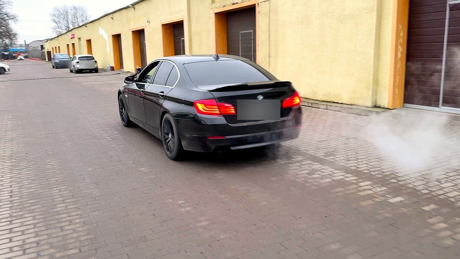 Чёрная BMW 5 F10 отъезжает от цеха «Выхлопухи» по брусчатке, из выхлопа идёт пар