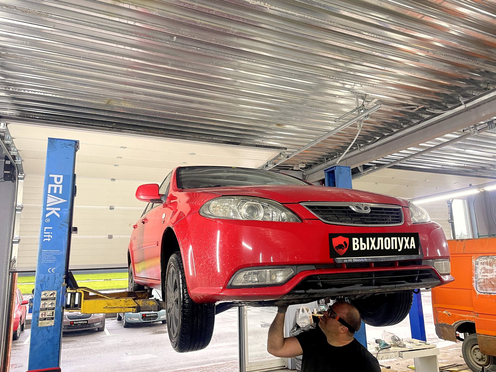 Красная Daewoo Gentra на подъёмнике в боксе «Выхлопухи» в Тельмана, вид спереди, мастер под машиной, номер закрыт плашкой