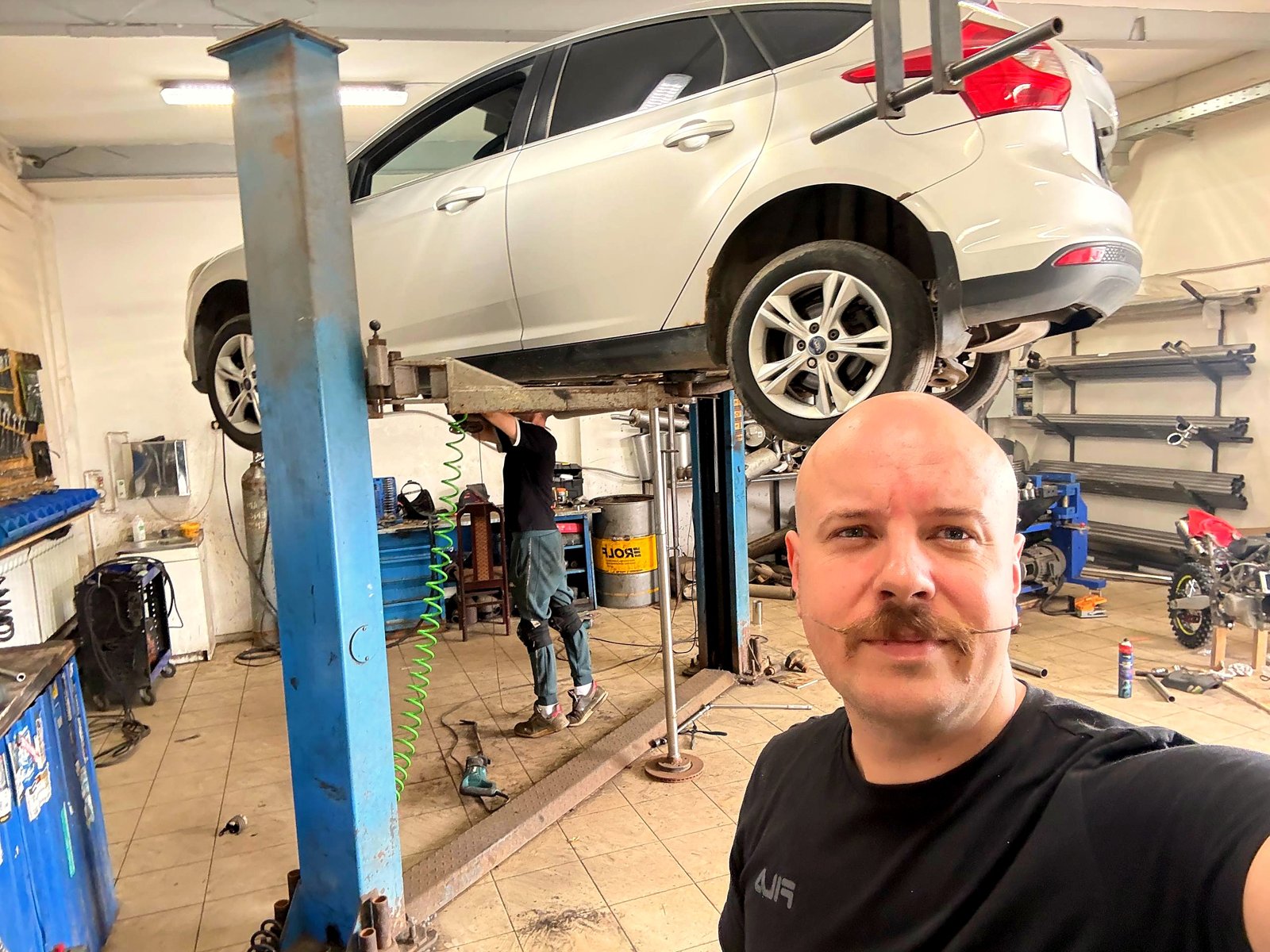 Белый Ford Focus 3 на подъёмнике в боксе «Выхлопухи», Роман на переднем плане, мастер работает под днищем