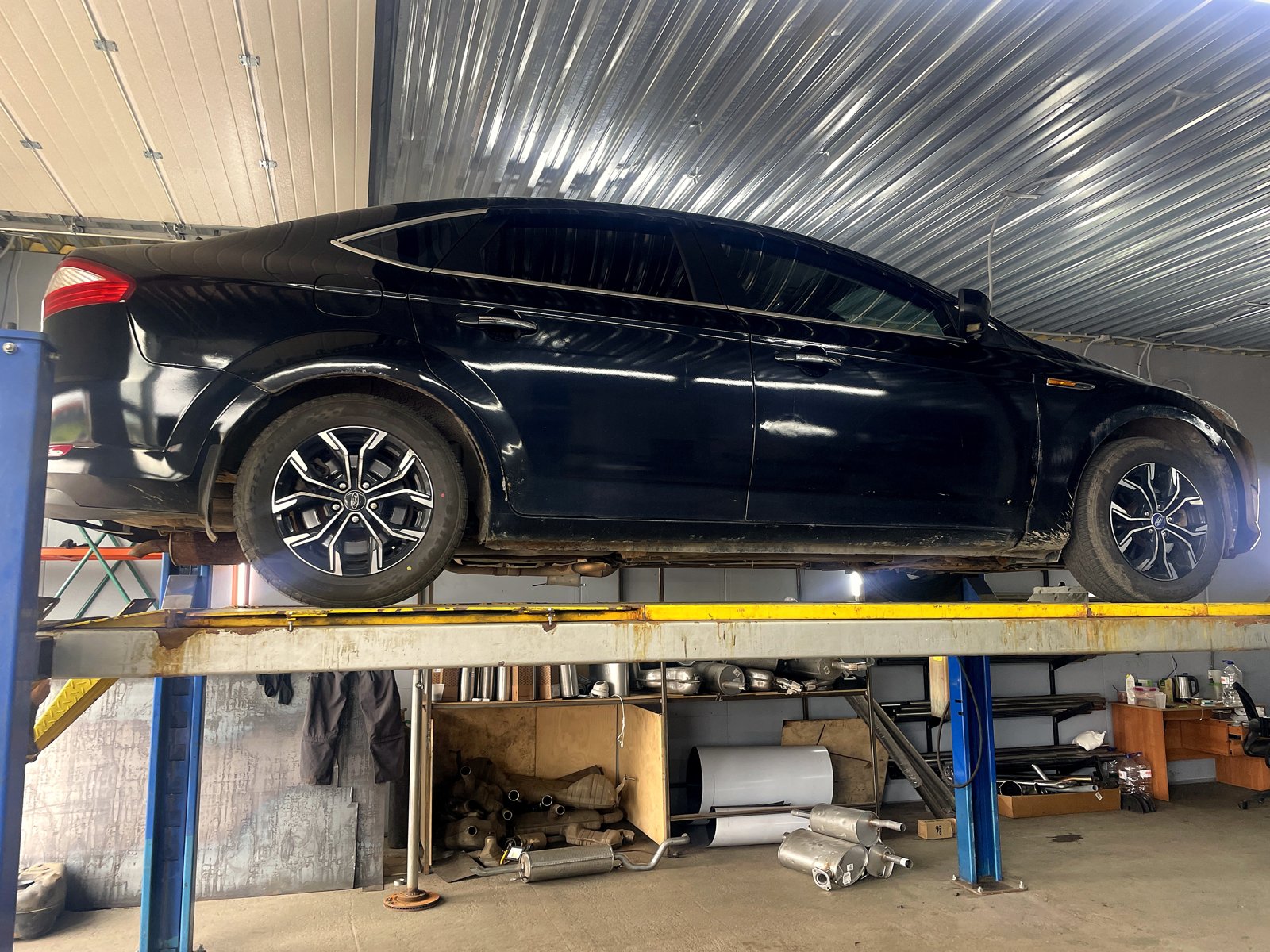 Чёрный Ford Mondeo 4 седан поднят на четырёхстоечный подъёмник в боксе «Выхлопухи», вид сбоку