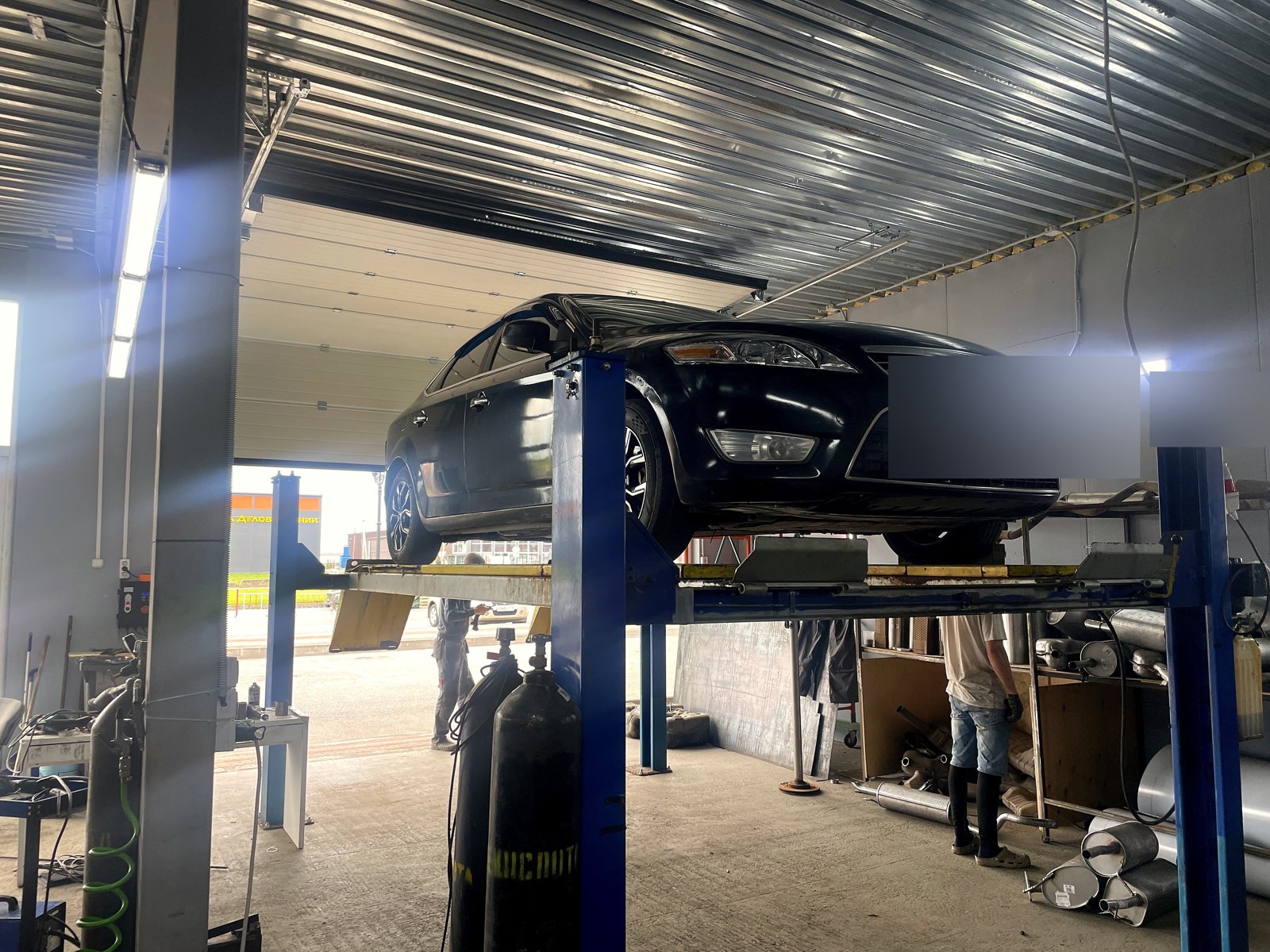 Ford Mondeo 4 на подъёмнике в цехе «Выхлопухи», общий план бокса со стеллажами заготовок