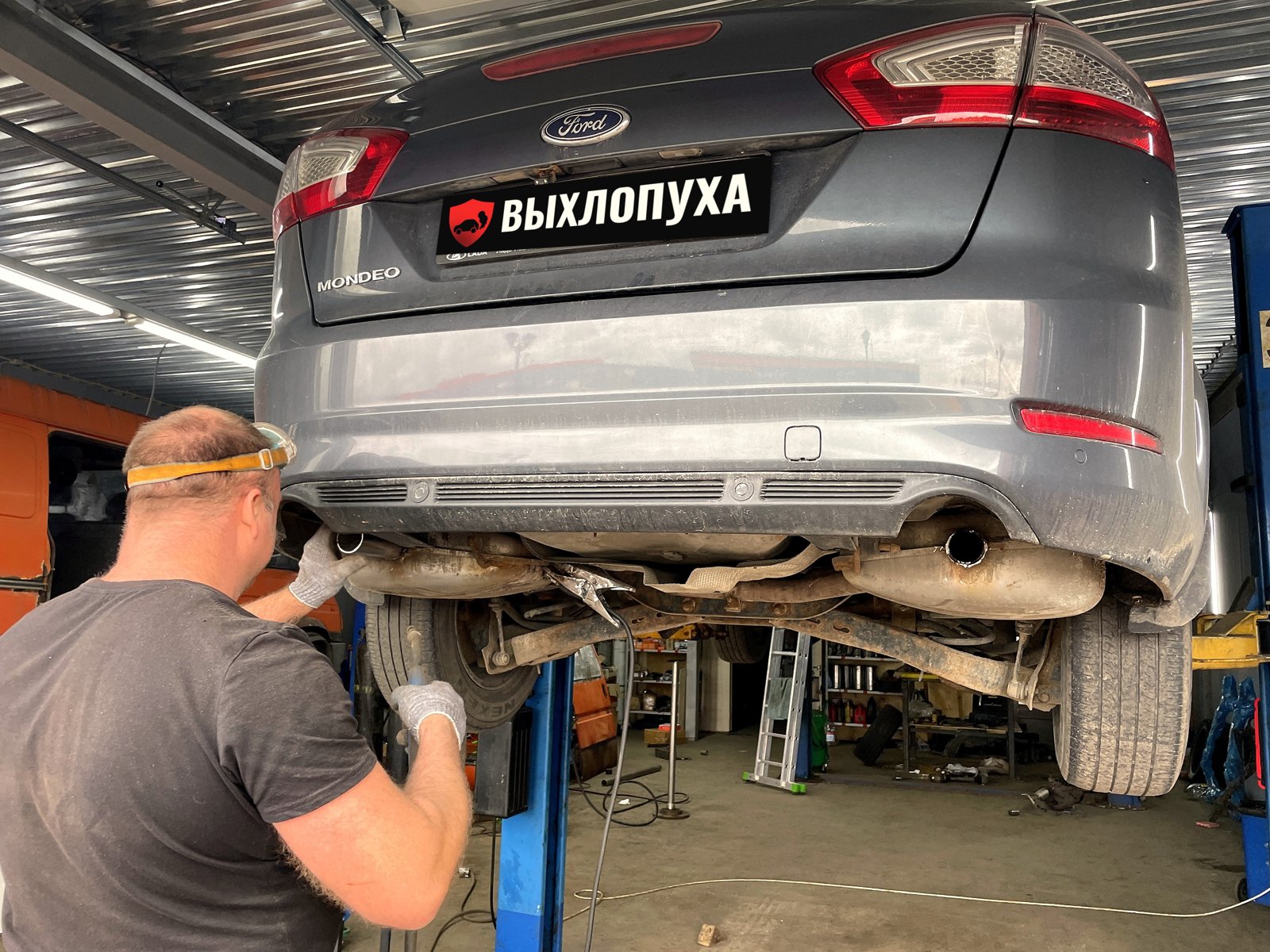 Мастер «Выхлопухи» у задних глушителей серого Ford Mondeo 4 на подъёмнике, штатный выход правого глушителя смотрит вниз