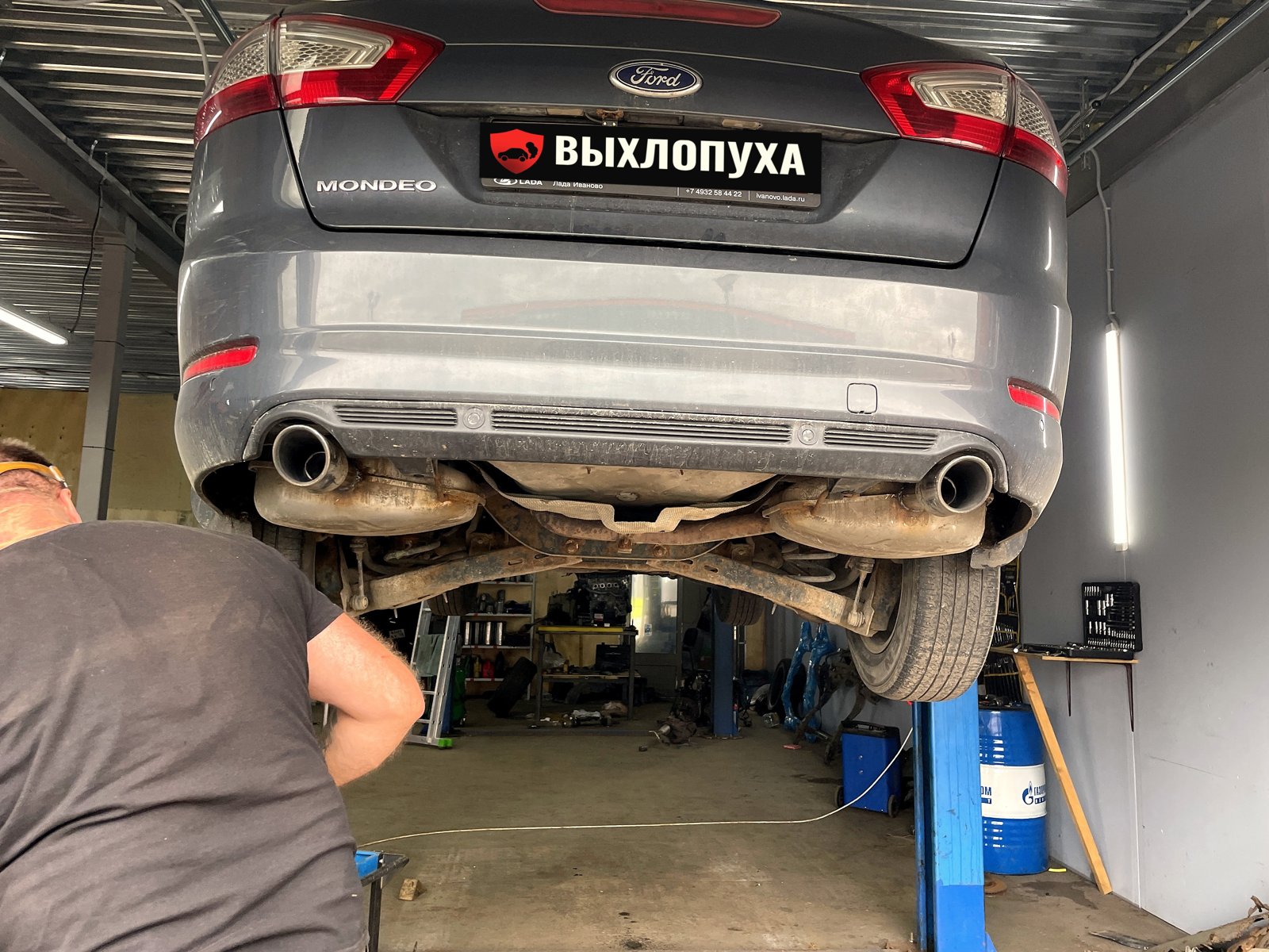 Серый Ford Mondeo 4 на подъёмнике, вид сзади: две насадки выхлопа смотрят назад из-под бампера, номер закрыт плашкой