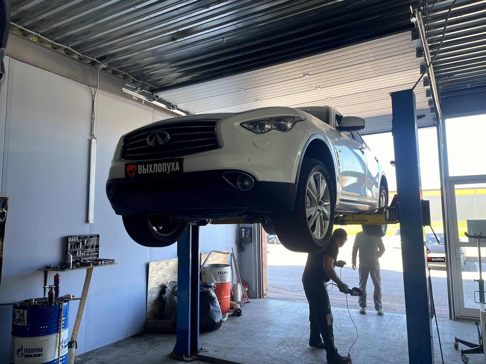 Белый Infiniti FX второго поколения на двухстоечном подъёмнике в цехе «Выхлопухи» на Тельмана, под машиной мастер с болгаркой