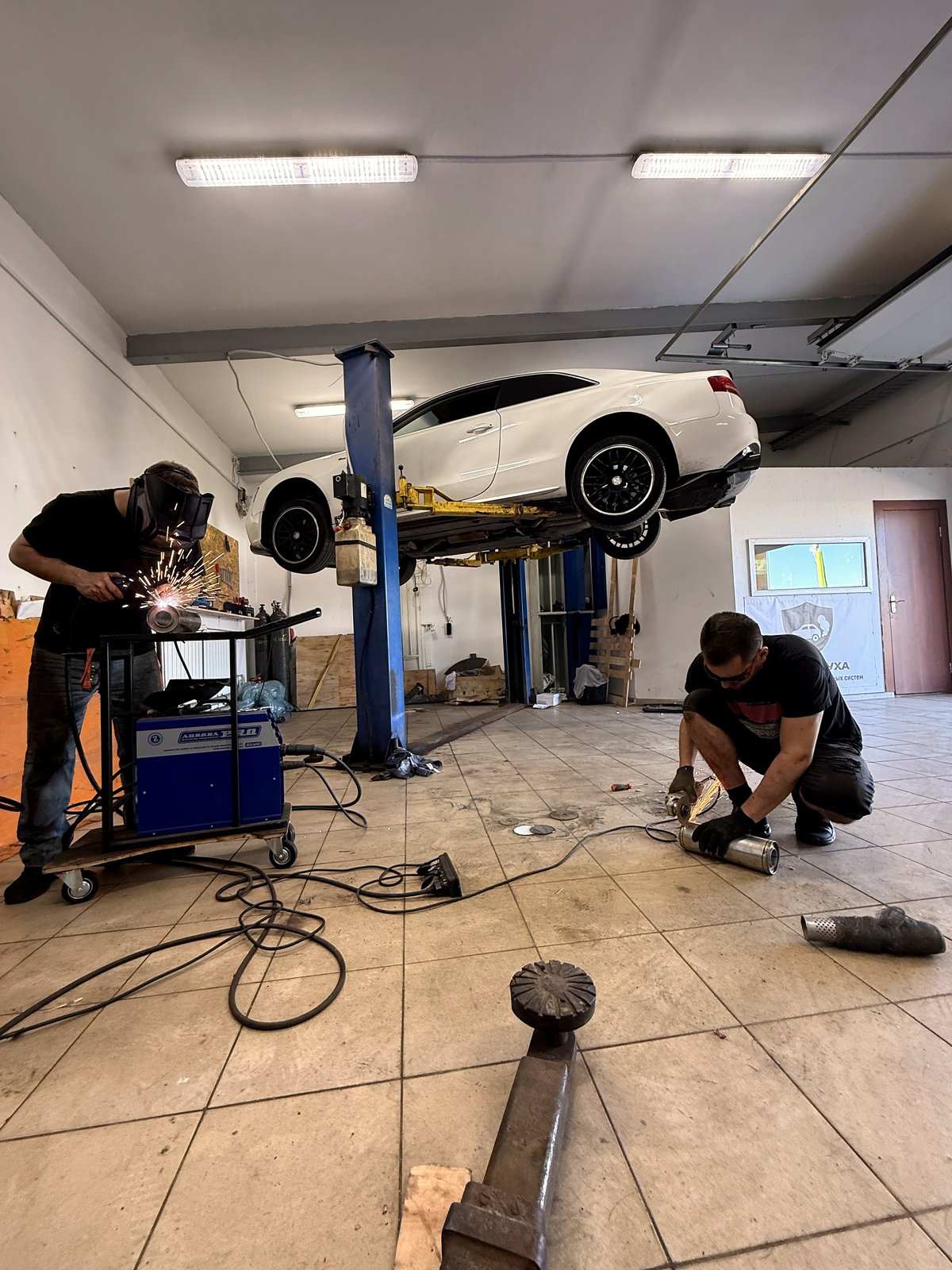 Белый Jaguar на подъёмнике в цеху «Выхлопухи», два мастера работают с выхлопной трассой