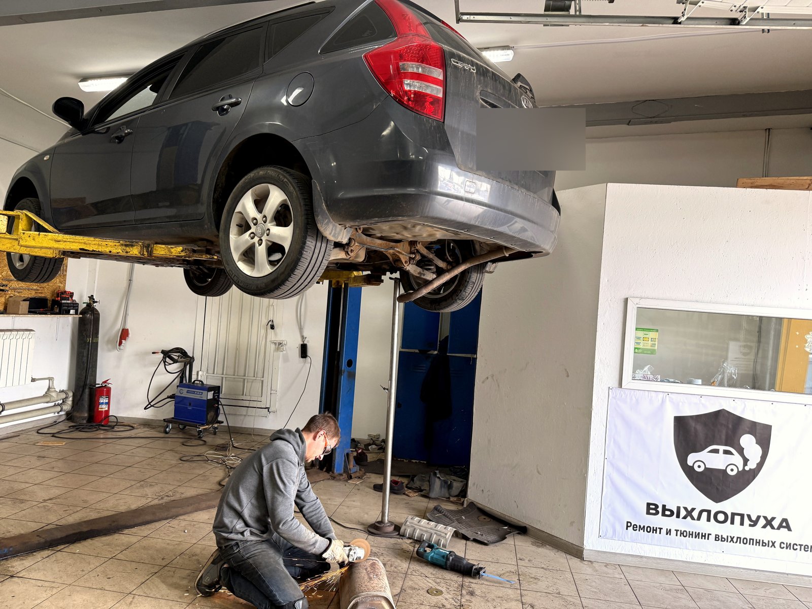 Серый Kia Ceed поднят на подъёмнике в цеху «Выхлопухи», мастер осматривает заднюю часть выхлопной трассы