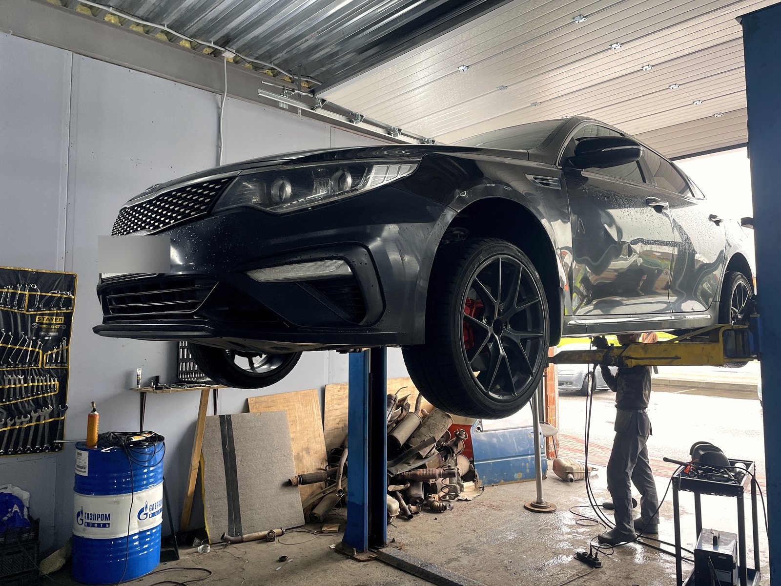 Тёмная Kia Optima на подъёмнике в боксе «Выхлопухи» в Тельмана, вид спереди, госномер замазан