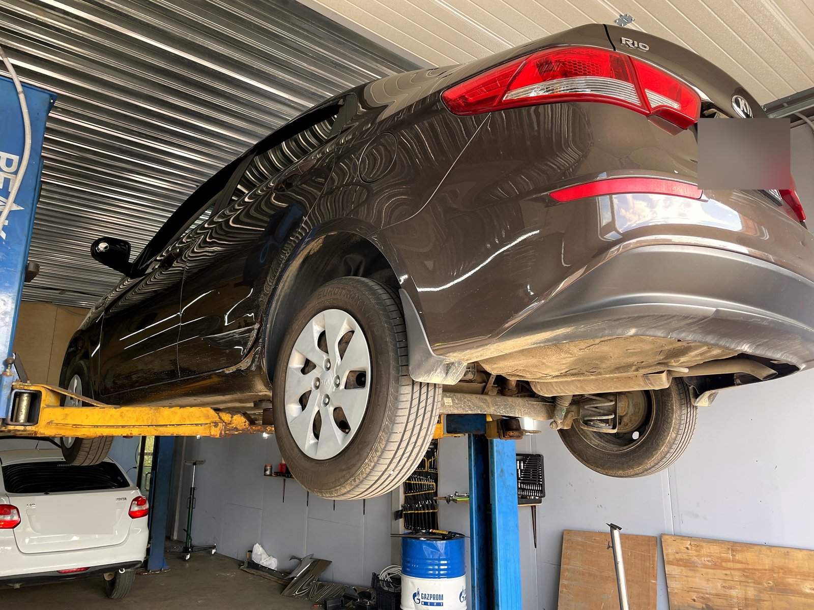 Задняя часть коричневого седана Kia Rio на подъёмнике в боксе «Выхлопухи», виден шильдик RIO и эмблема Kia, номера замазаны