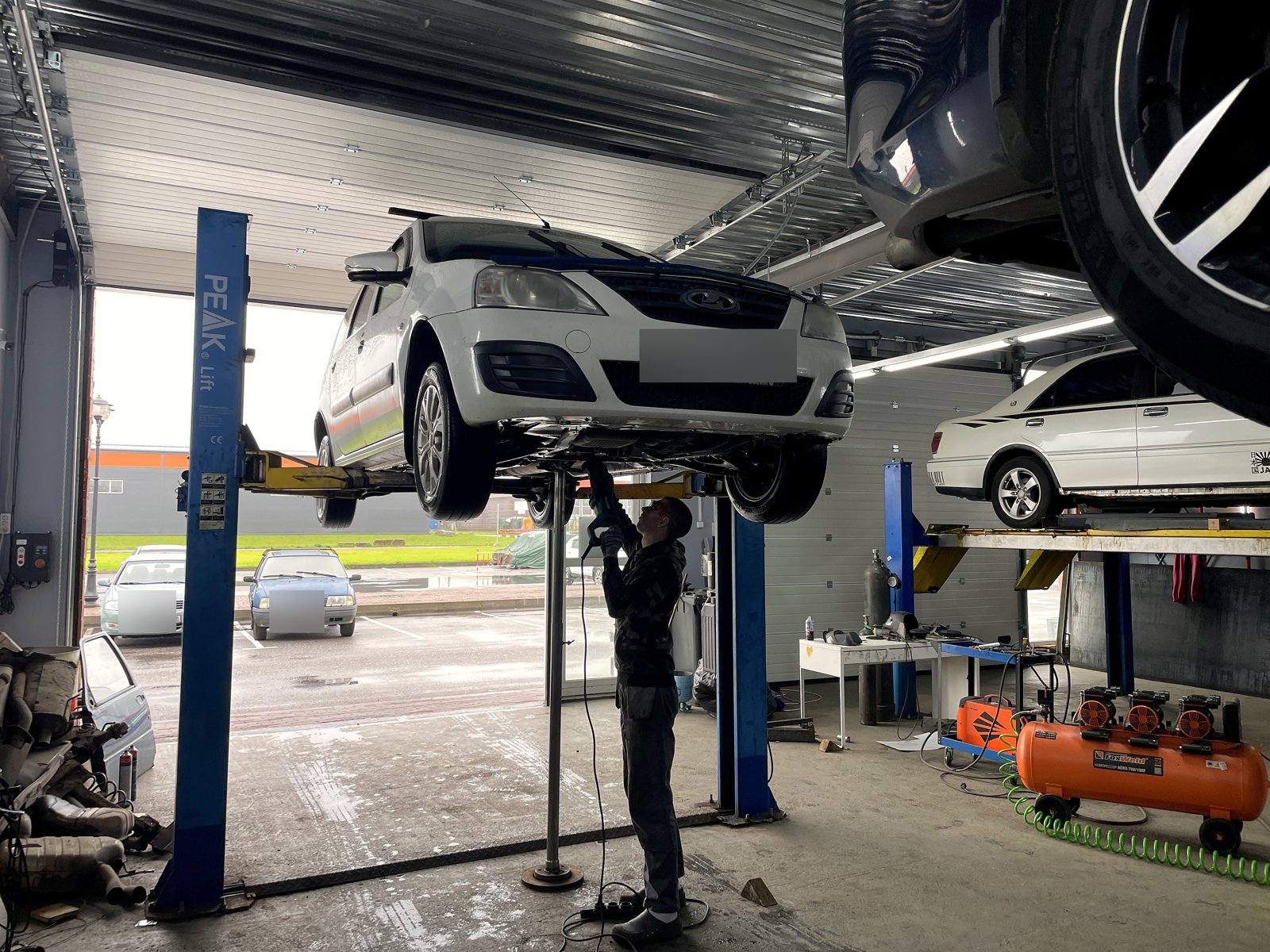 Белая Lada Largus на подъёмнике в боксе «Выхлопухи» в Тельмана, госномер замазан