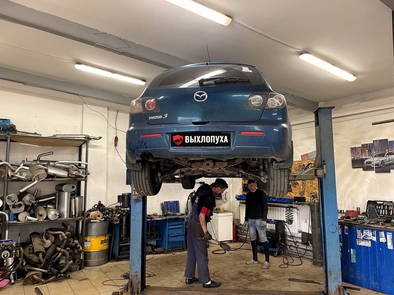 Синяя Mazda 3 хэтчбек на двухстоечном подъёмнике в цехе «Выхлопухи» на Южном шоссе, под машиной двое мастеров