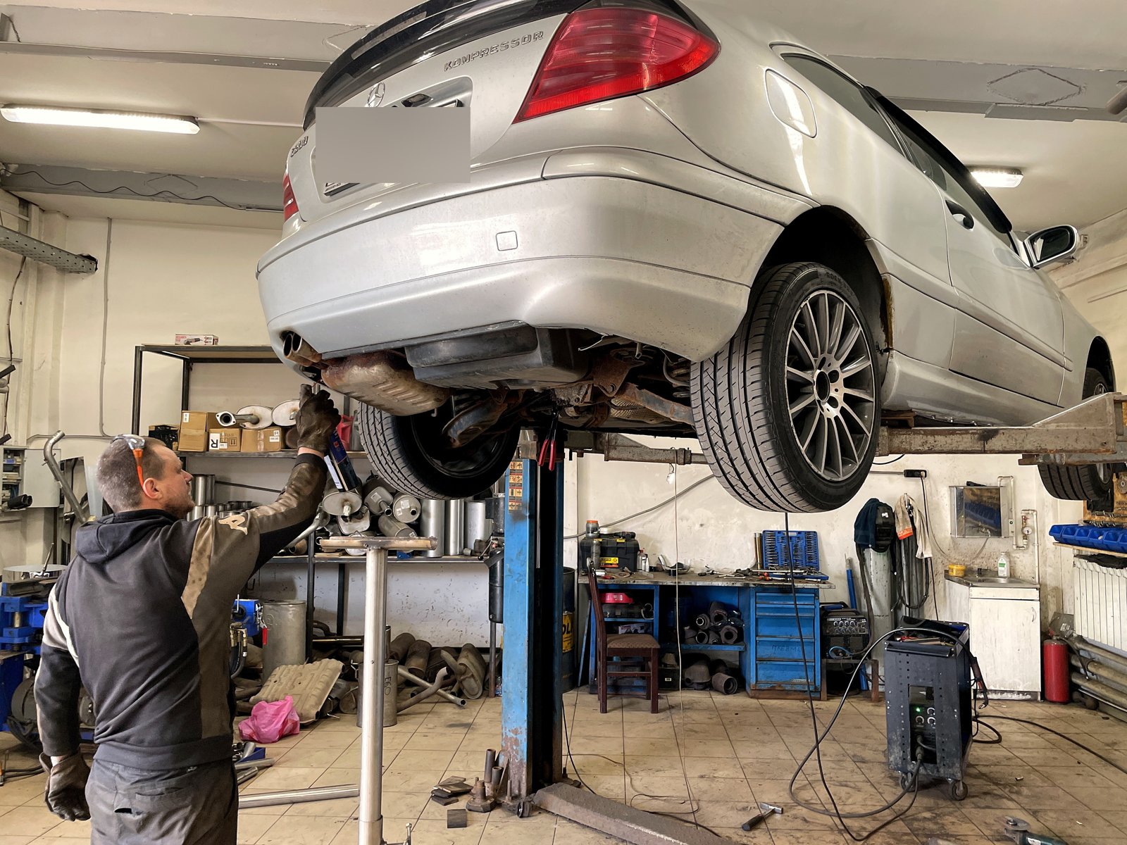 Серебристый Mercedes C200 Kompressor в кузове купе поднят на двухстоечном подъёмнике в боксе «Выхлопухи», вид сзади