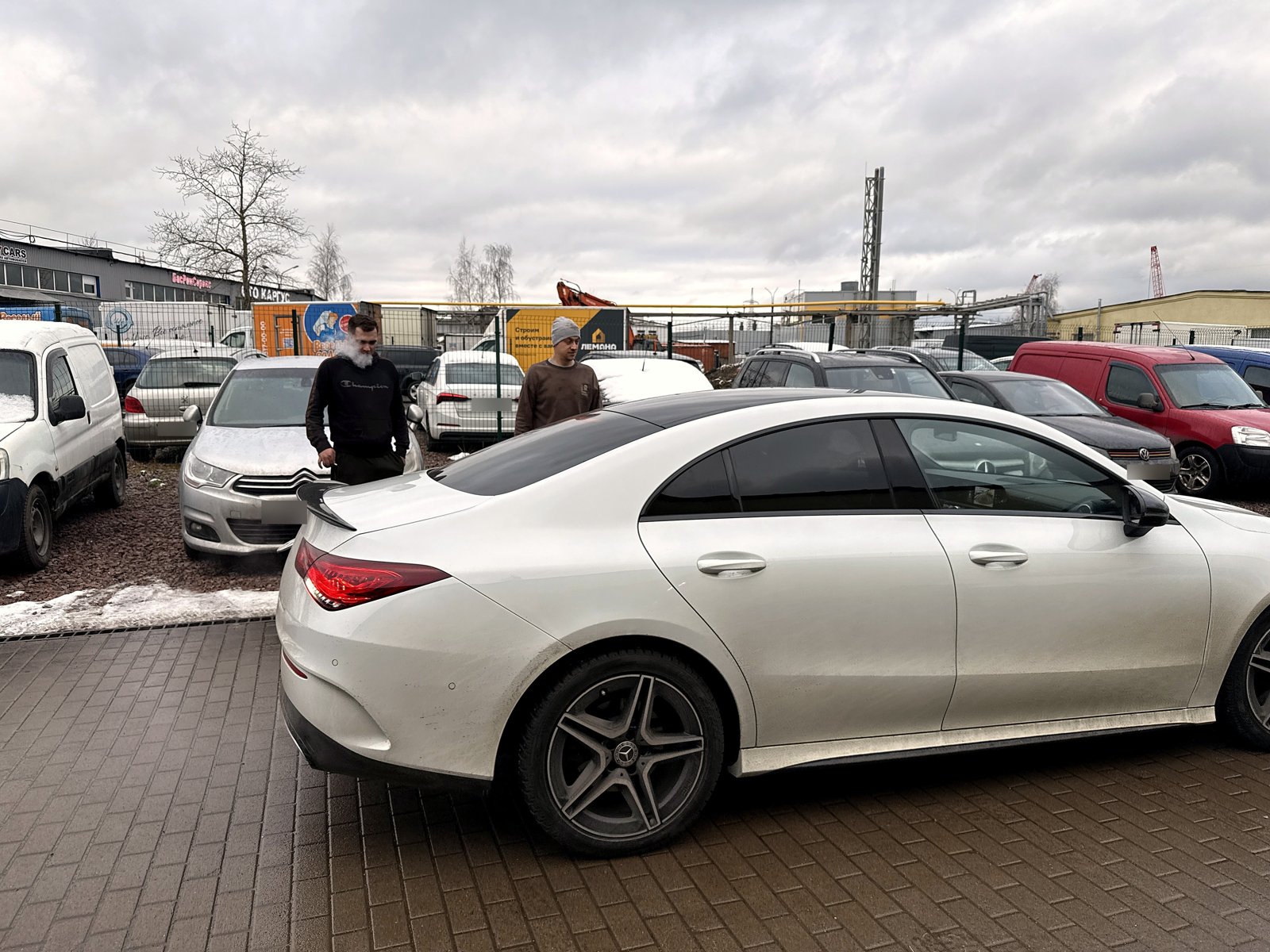 Белый Mercedes CLA на площадке у цеха «Выхлопухи» после работы, вид сзади сбоку, рядом стоят двое
