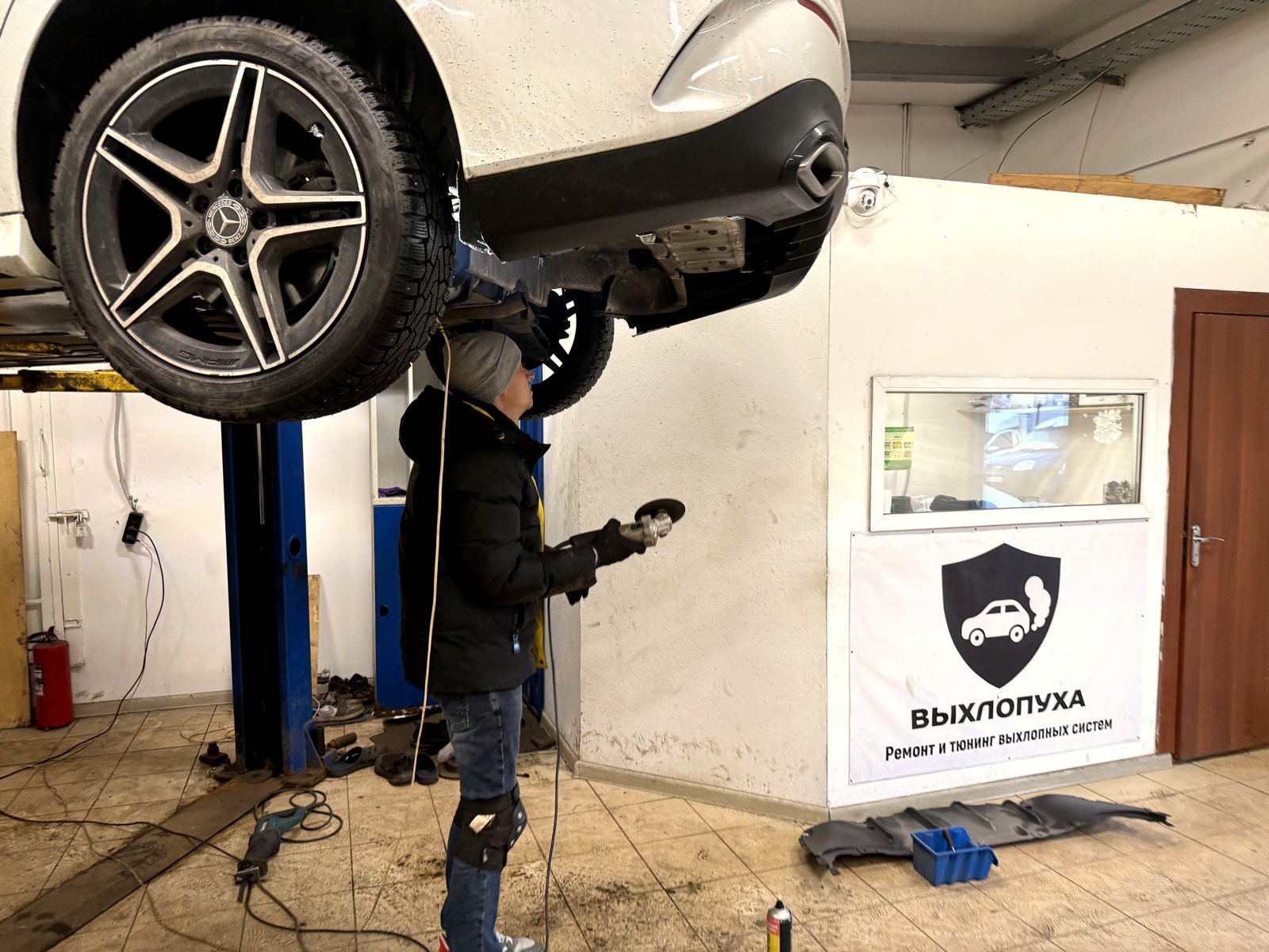 Белый Mercedes CLA на подъёмнике в цеху «Выхлопухи», мастер под задней частью с болгаркой