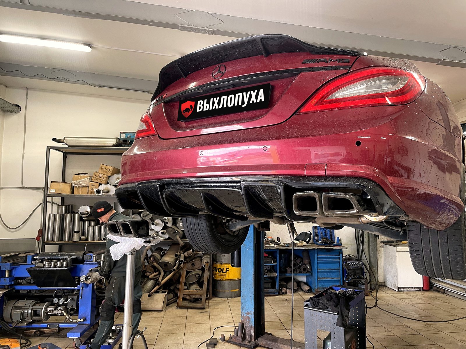 Бордовый Mercedes CLS C218 в обвесе AMG на подъёмнике в боксе «Выхлопухи» на Южном шоссе, под диффузором четыре насадки, номер закрыт плашкой