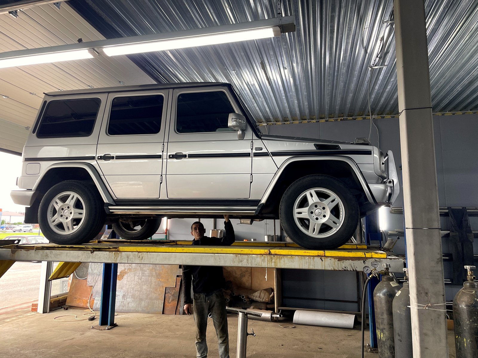 Серебристый Mercedes-Benz G-класс W463 на подъёмнике в боксе «Выхлопухи» в Тельмана, вид сбоку, под машиной мастер