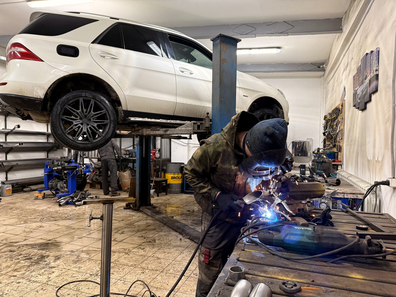 Белый Mercedes ML 350 на подъёмнике в цеху «Выхлопухи», рядом мастер варит на верстаке