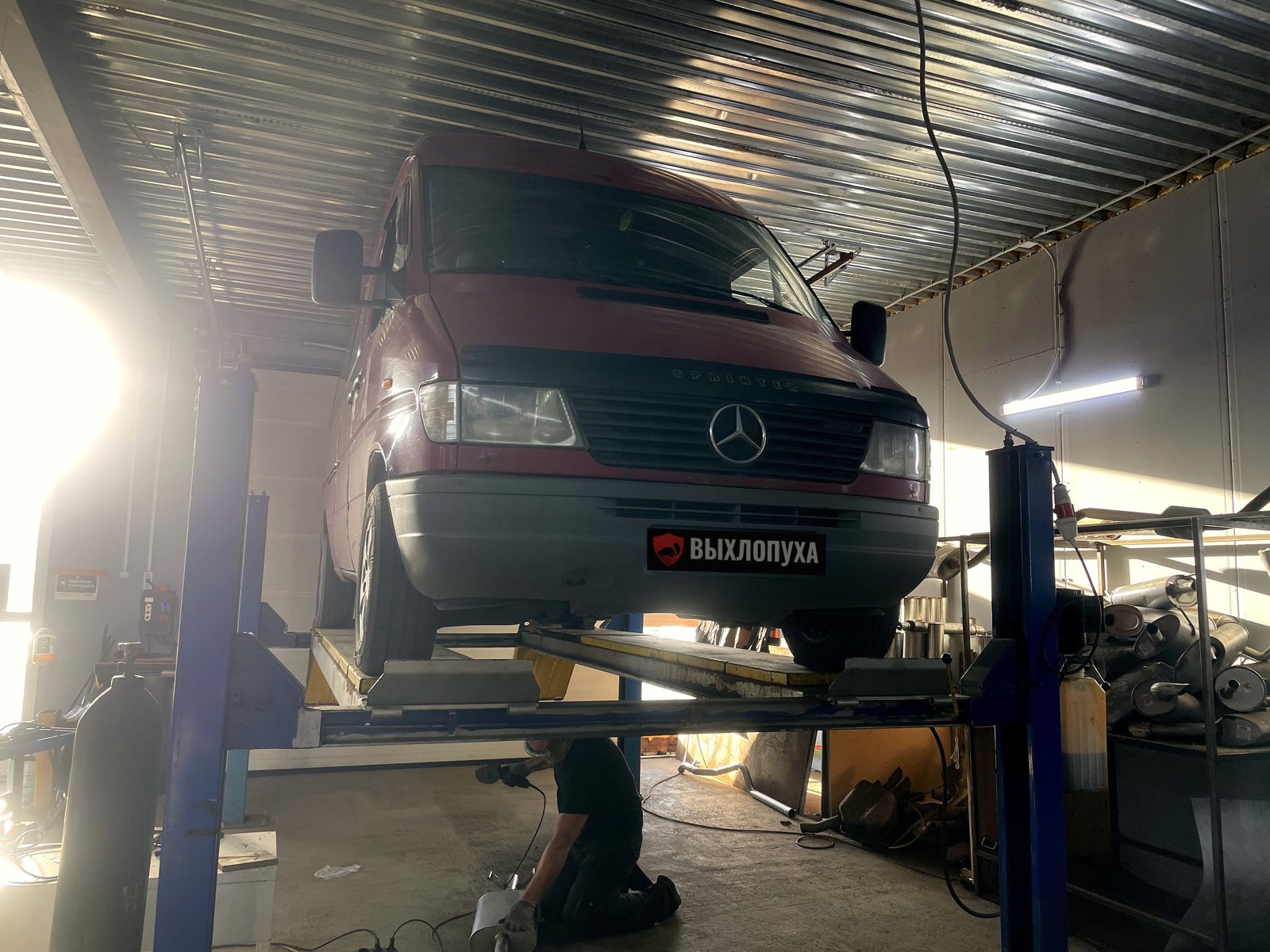 Бордовый фургон Mercedes-Benz Sprinter первого поколения на подъёмнике в боксе «Выхлопухи» в Тельмана, вид спереди, номер закрыт плашкой
