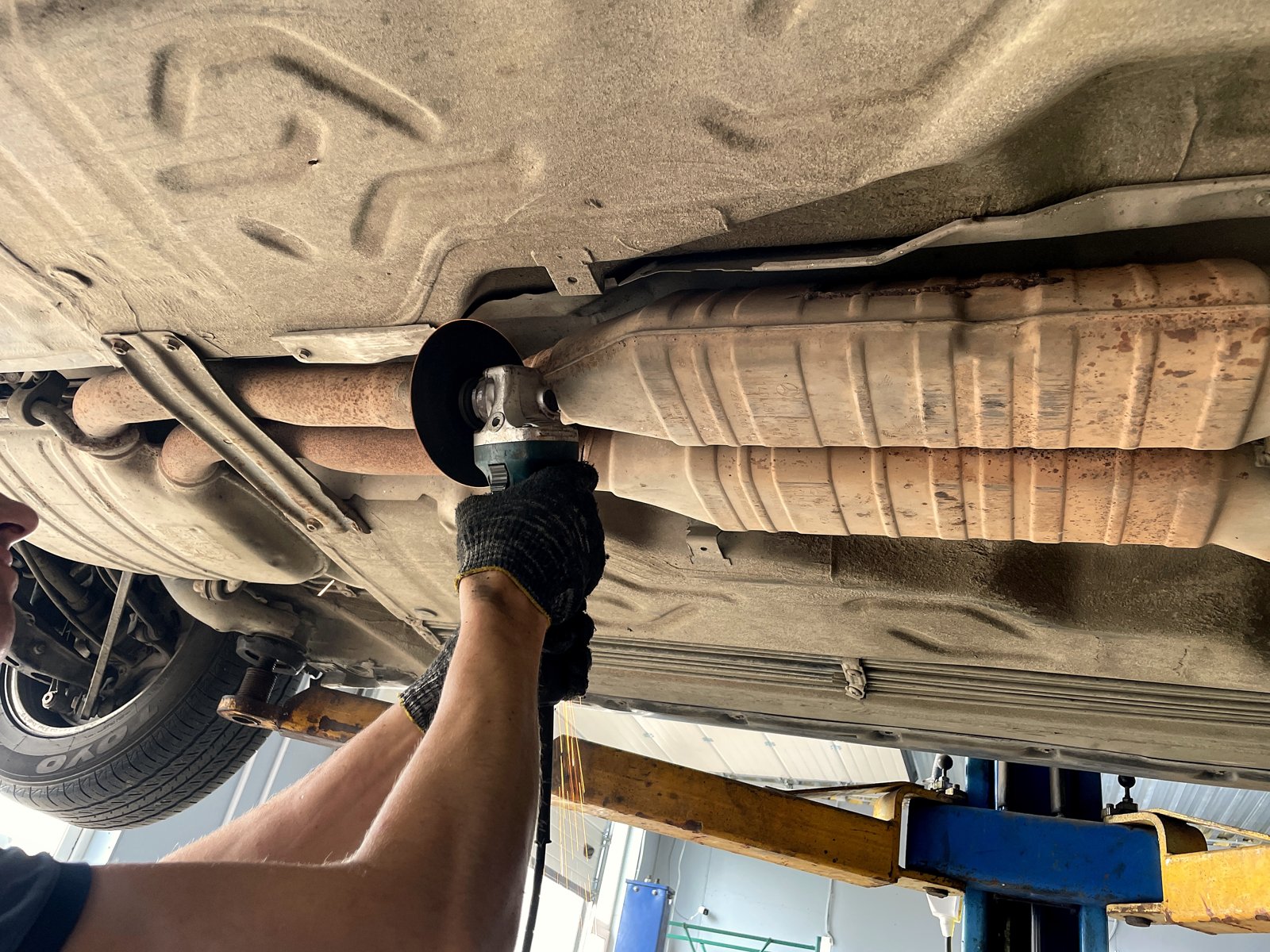 Старый овальный корпус под днищем Mercedes W140, белёсый металл с рыжими пятнами, мастер подводит болгарку