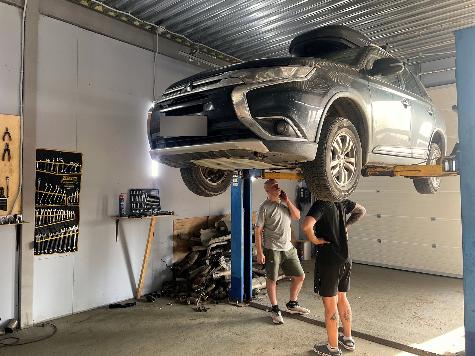 Тёмный Mitsubishi Outlander поднят на двухстоечном подъёмнике в боксе «Выхлопухи», мастера осматривают машину снизу
