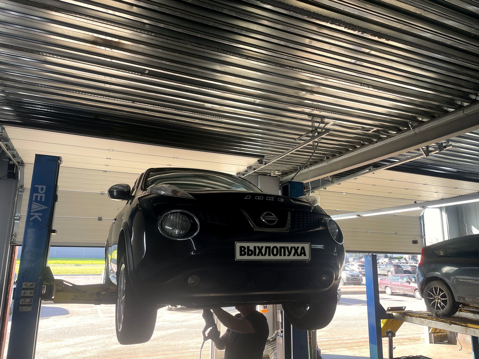 Чёрный Nissan Juke первого поколения поднят на двухстоечном подъёмнике в боксе «Выхлопухи» в Тельмана, вид спереди, под машиной мастер, номер закрыт плашкой