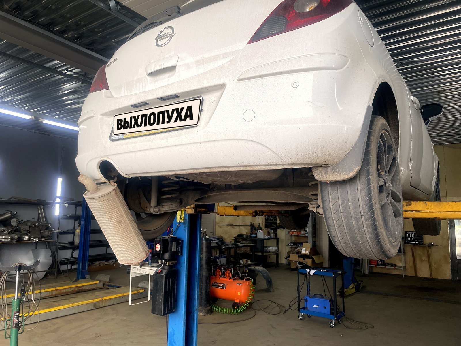 Корма белого Opel Corsa D на подъёмнике снизу, задняя банка глушителя опущена и висит, номер закрыт плашкой