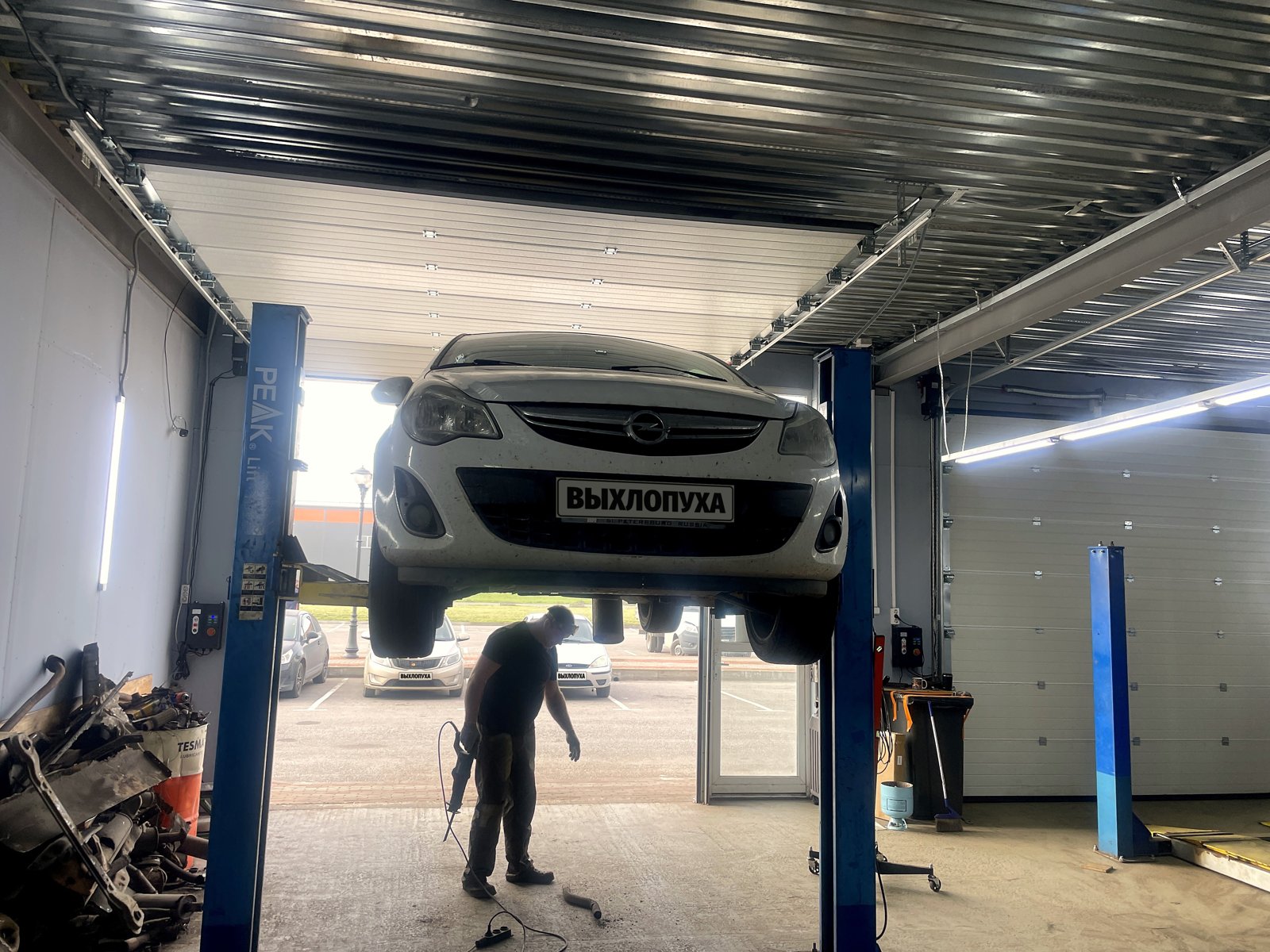 Белый Opel Corsa D поднят на двухстоечном подъёмнике в боксе «Выхлопухи» в Тельмана, вид спереди, под машиной мастер с пилой, номера закрыты плашками