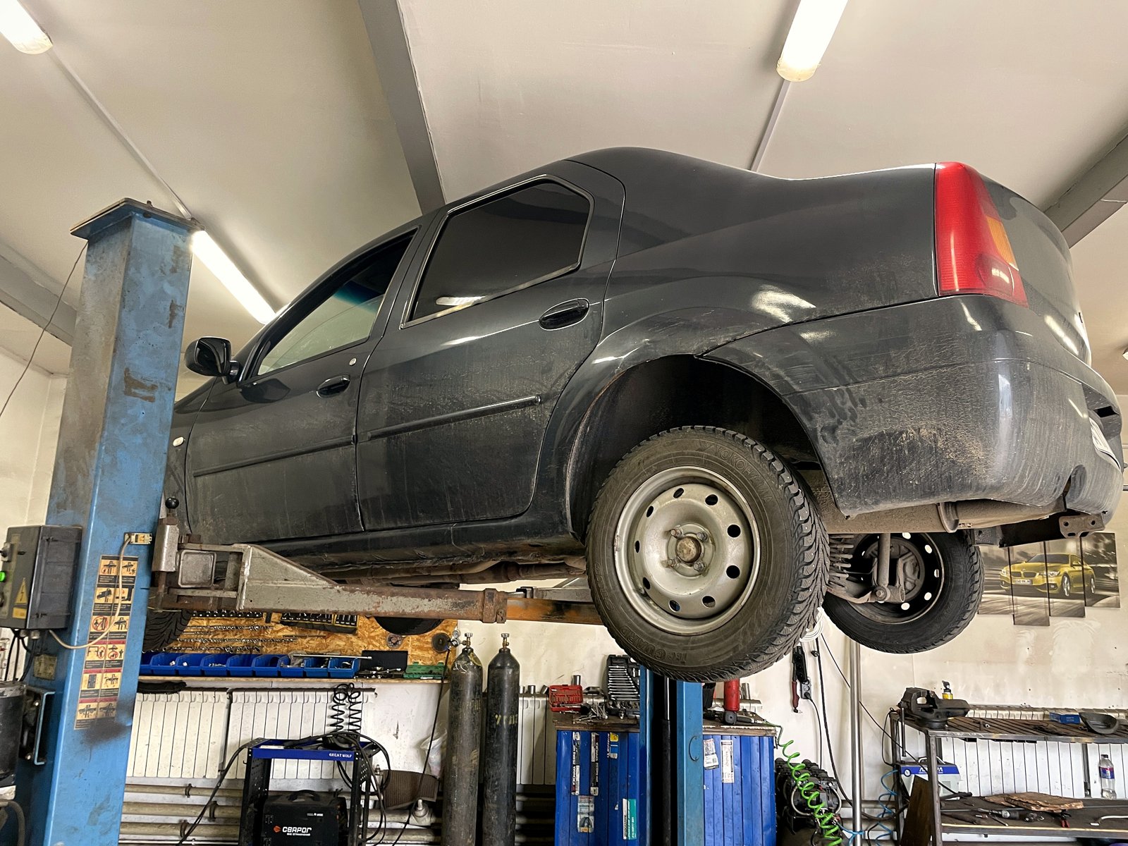 Тёмный Renault Logan первого поколения на двухстоечном подъёмнике в цехе «Выхлопухи» на Южном шоссе