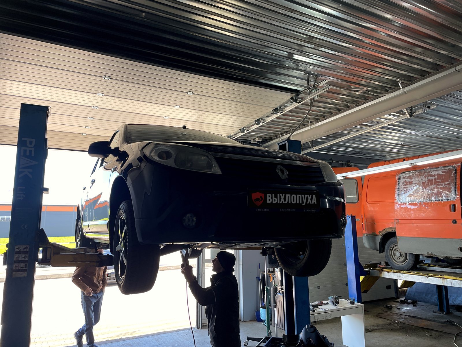 Тёмный Renault Sandero первого поколения на подъёмнике в цехе «Выхлопухи» на Тельмана, мастер под машиной