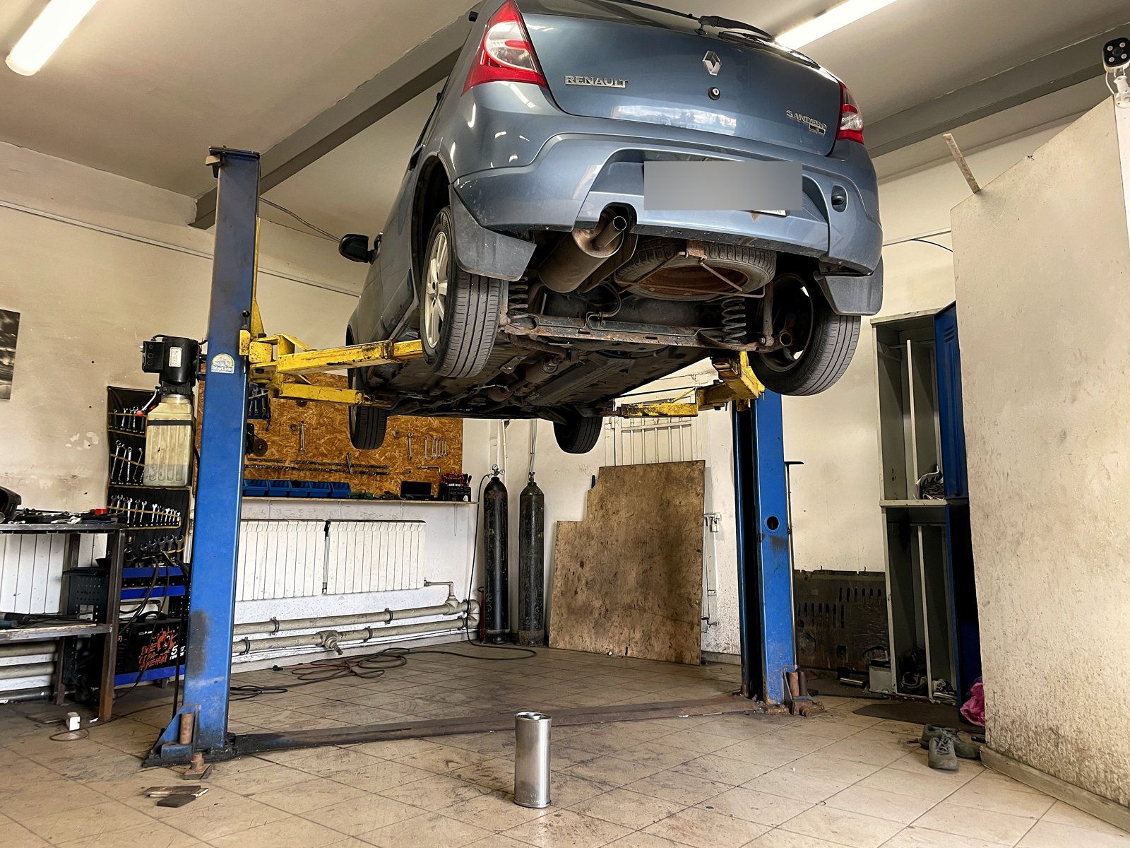Серо-голубой Renault Sandero поднят на двухстоечном подъёмнике в боксе «Выхлопухи», вид сзади