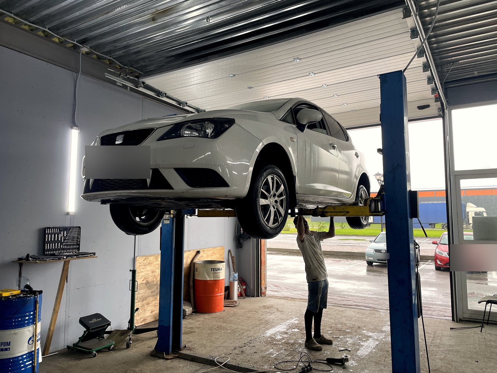 Белый SEAT Ibiza поднят на двухстоечном подъёмнике в боксе «Выхлопухи», мастер работает снизу, передний номер замазан