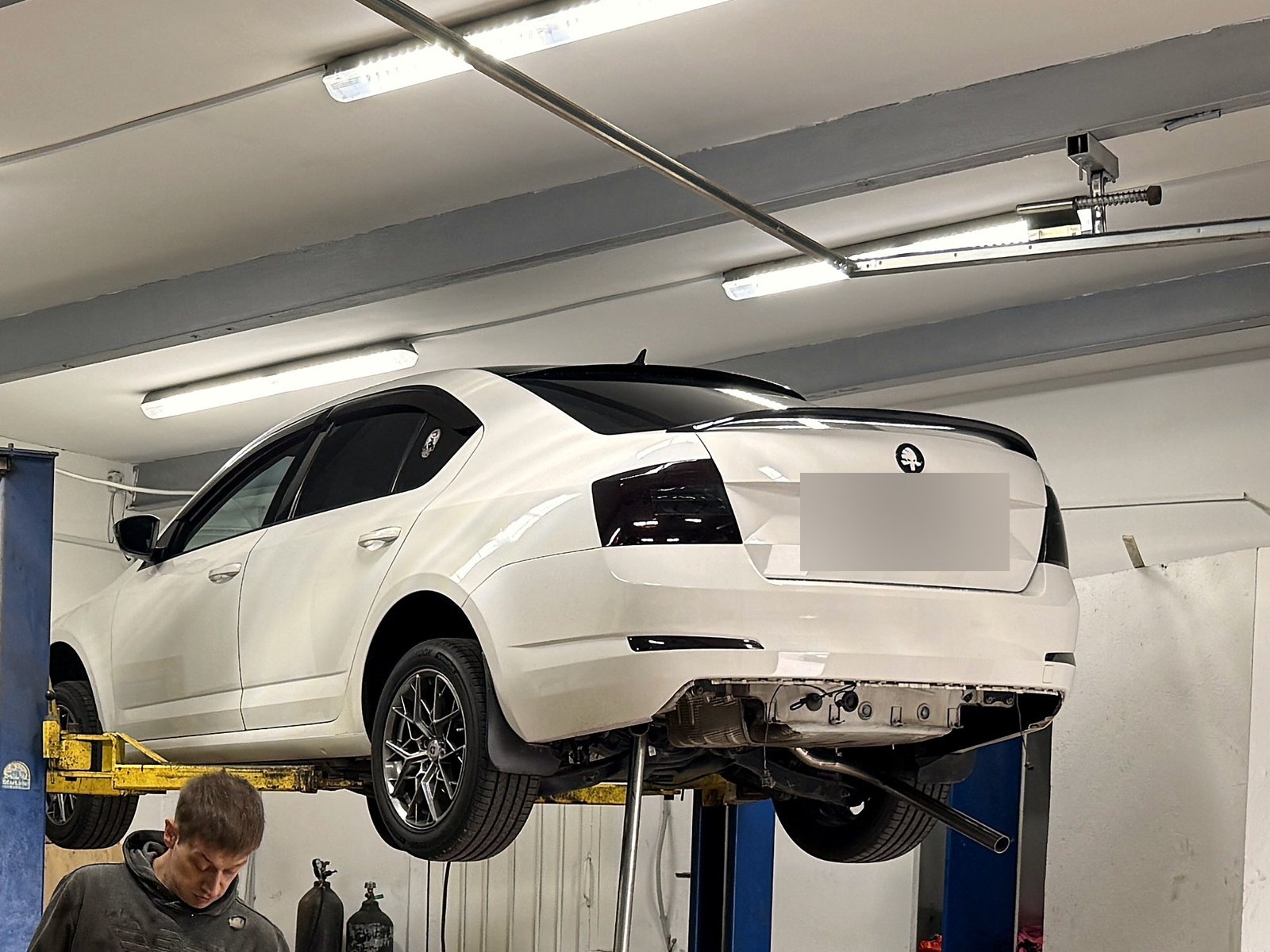 Белая Skoda Octavia целиком на подъёмнике в цеху «Выхлопухи»: тонированные фонари, чёрный спойлер, выхлоп ещё выходит одной трубой сбоку