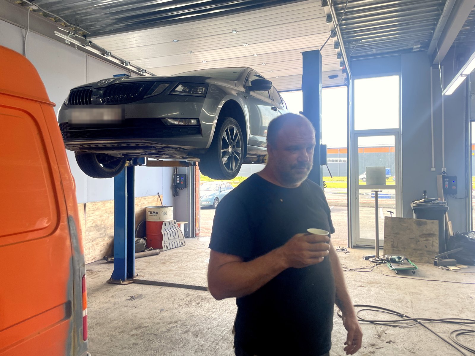 Серый лифтбек Skoda Octavia поднят на подъёмнике в боксе «Выхлопухи», рядом Роман с кофе