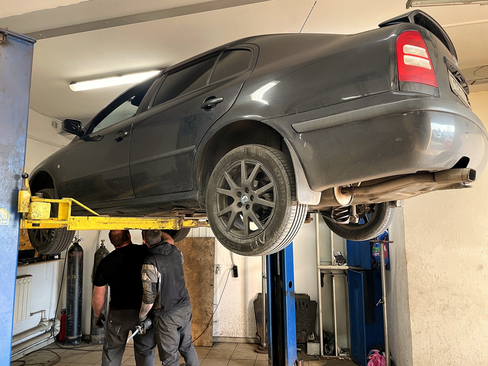 Тёмно-серая Škoda Octavia первого поколения поднята на двухстоечном подъёмнике в боксе «Выхлопухи», вид сзади-сбоку