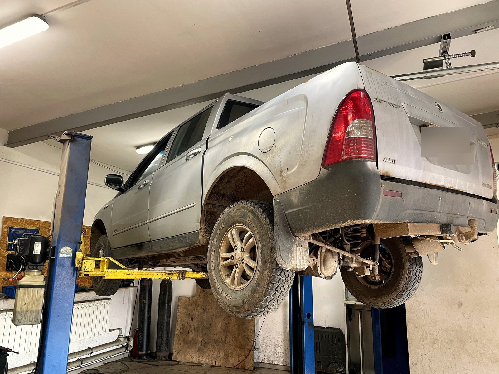 Серебристый пикап SsangYong Actyon Sports поднят на подъёмнике в боксе «Выхлопухи», вид сзади, номер замазан