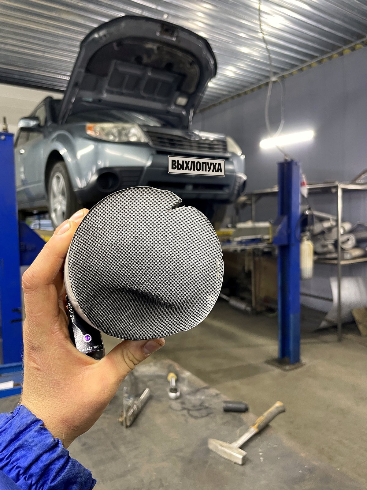 Вырезанная сота катализатора в руке, на фоне Subaru Forester на подъёмнике