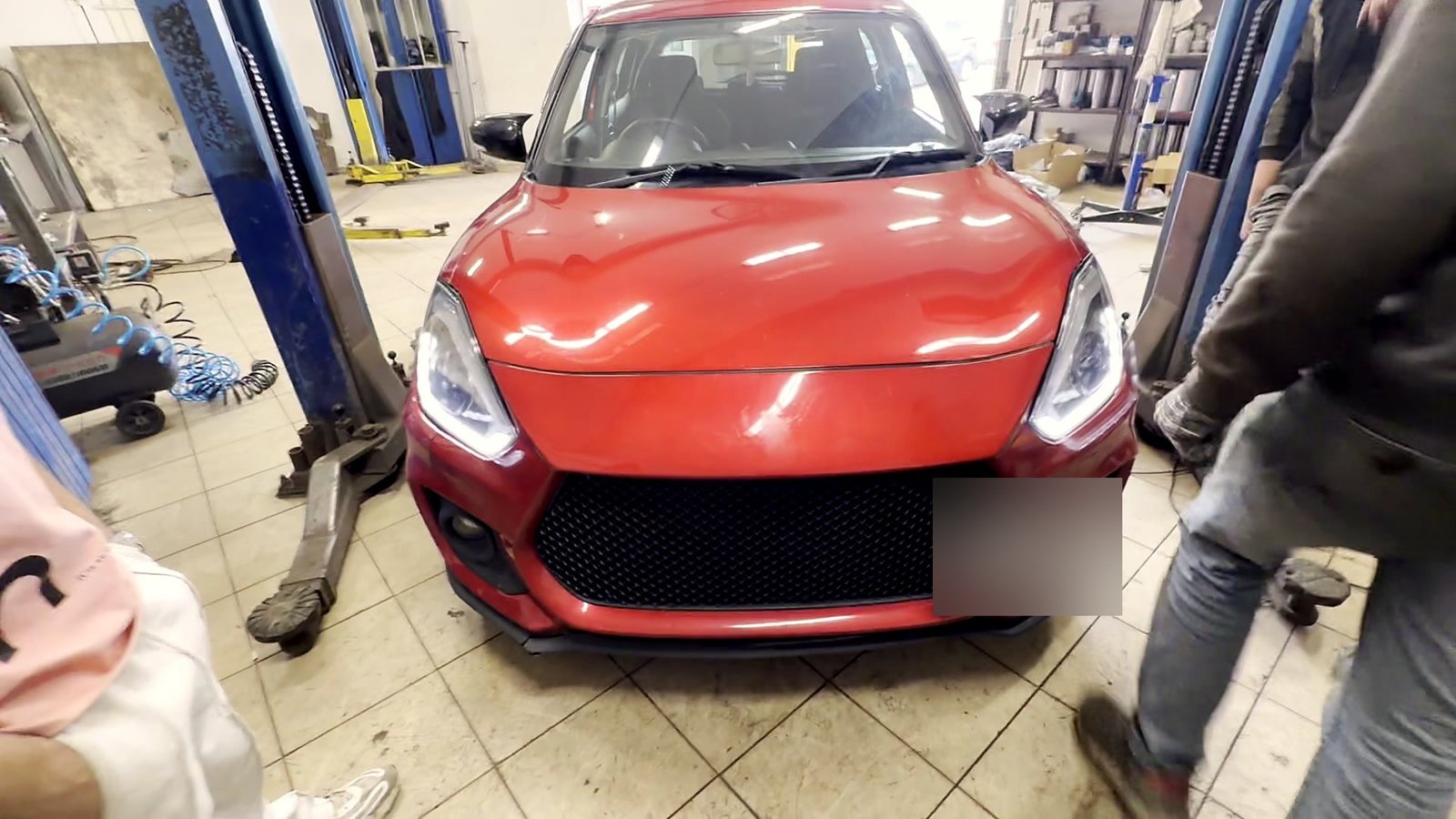 Красный Suzuki Swift в цеху «Выхлопухи» между стойками подъёмника, вид спереди