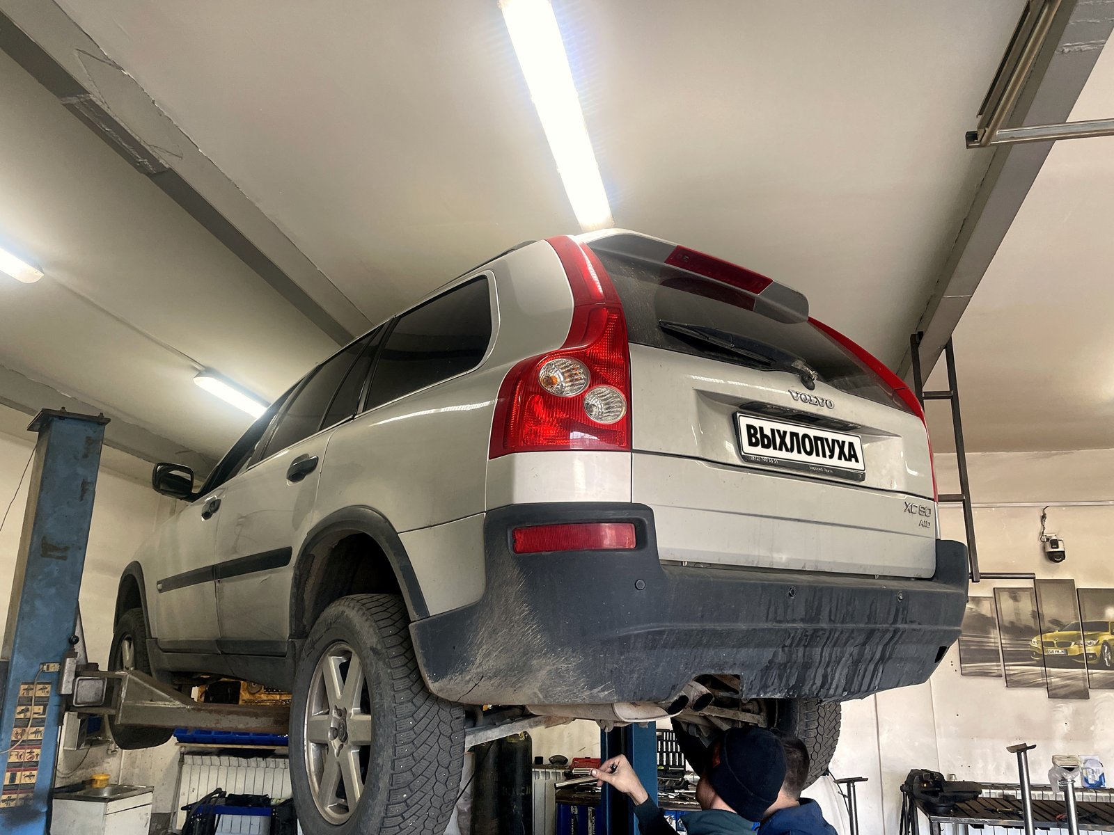 Серебристый Volvo XC90 первого поколения на подъёмнике в боксе «Выхлопухи» на Южном шоссе, вид сзади, номер закрыт плашкой
