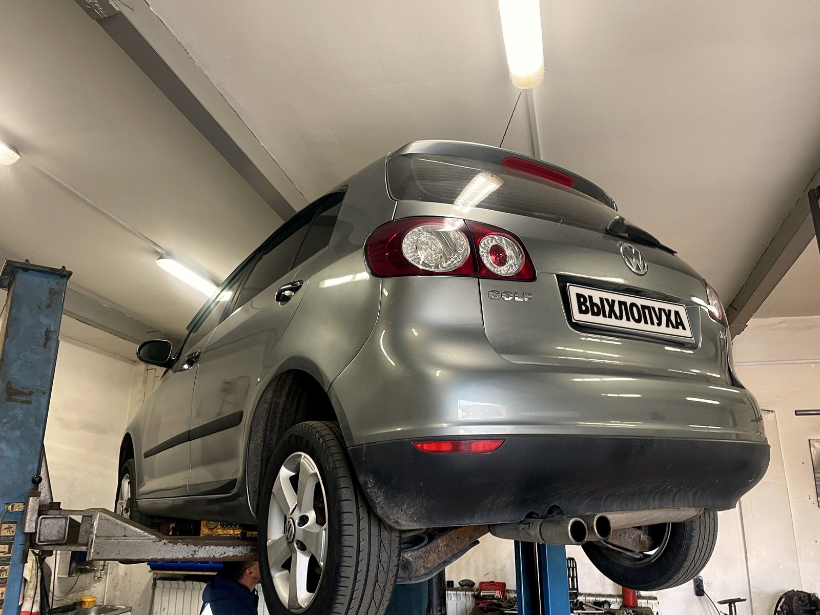 Серый VW Golf Plus поднят на подъёмнике в боксе «Выхлопухи» на Южном шоссе, вид сзади снизу, номер закрыт плашкой