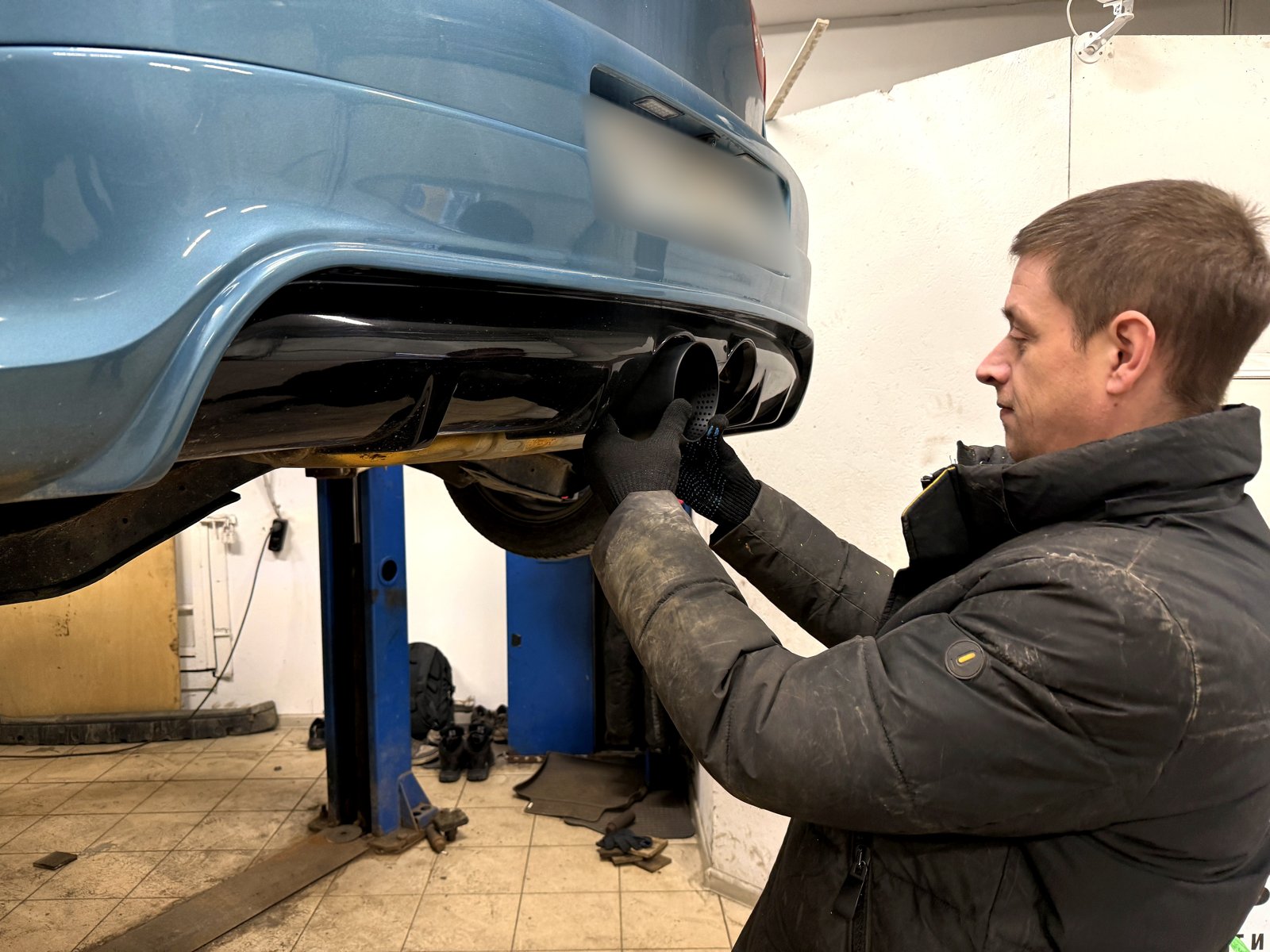 Мастер «Выхлопухи» ставит чёрную насадку в вырез диффузора голубого VW Golf на подъёмнике