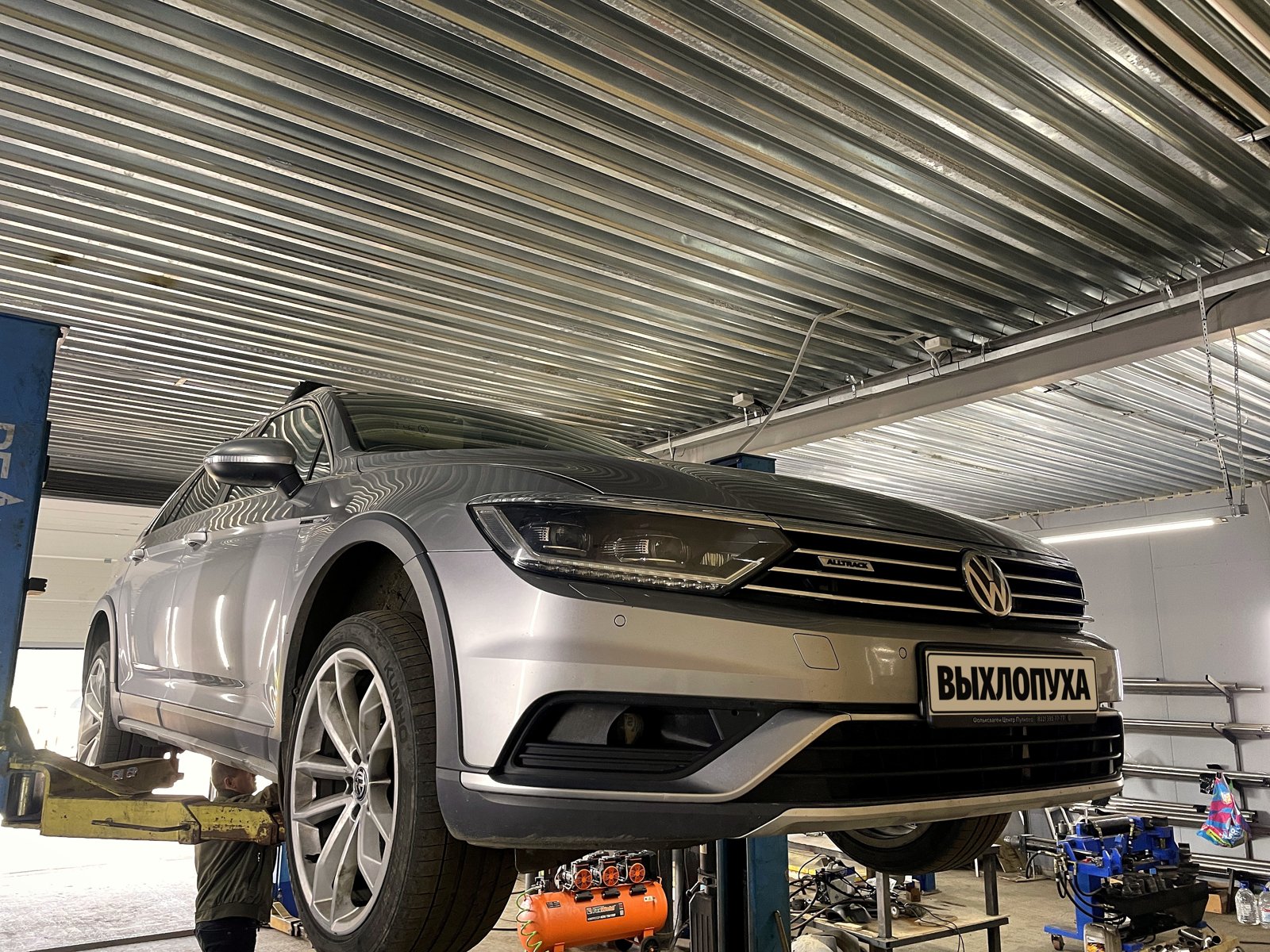 Серебристый VW Passat Alltrack поднят на двухстоечном подъёмнике в боксе «Выхлопухи» в Тельмана, вид спереди, номер закрыт плашкой