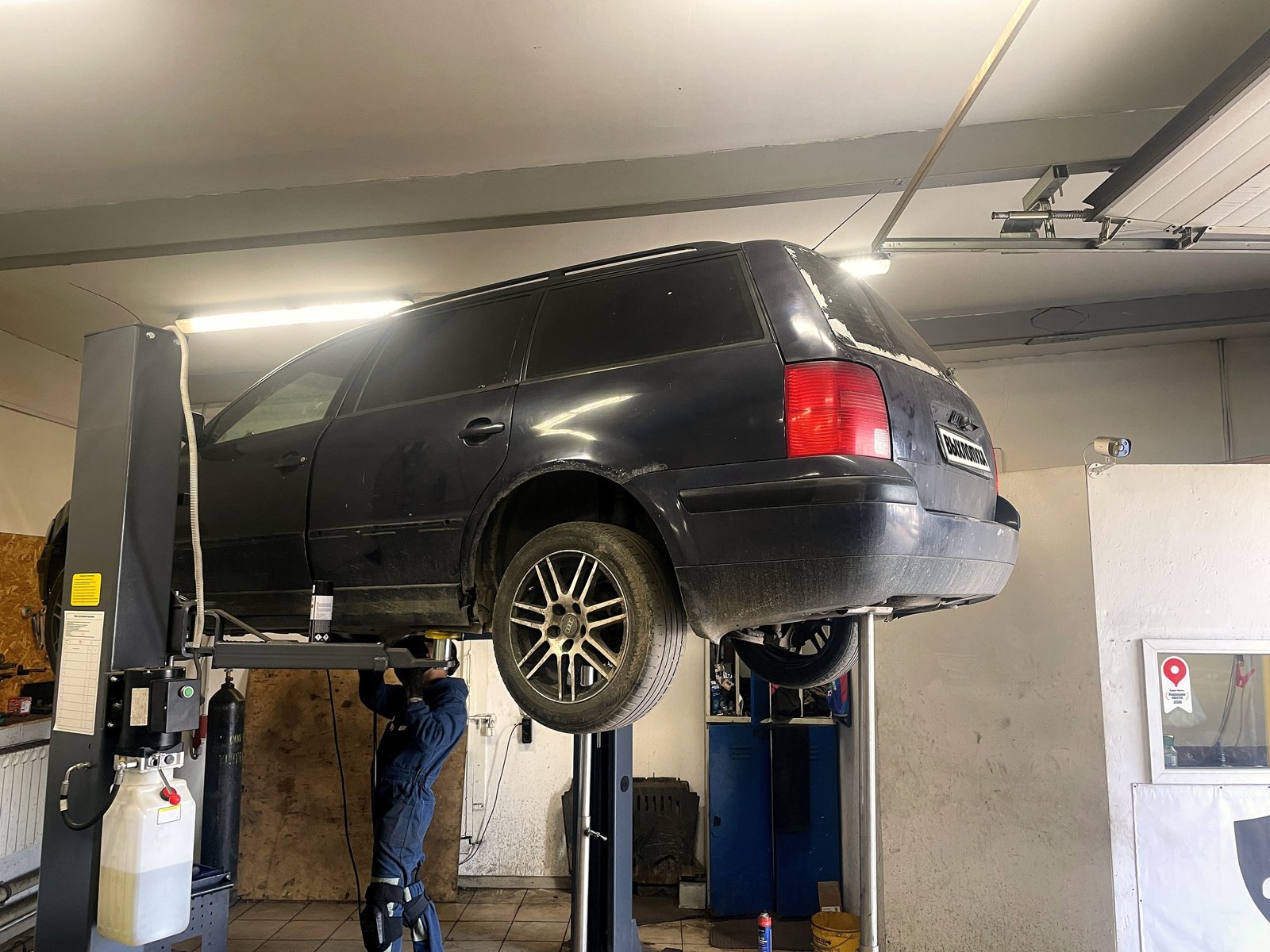 Чёрный VW Passat B5 универсал поднят на подъёмнике в боксе «Выхлопухи» на Южном шоссе, вид сбоку с кормы, номер закрыт плашкой