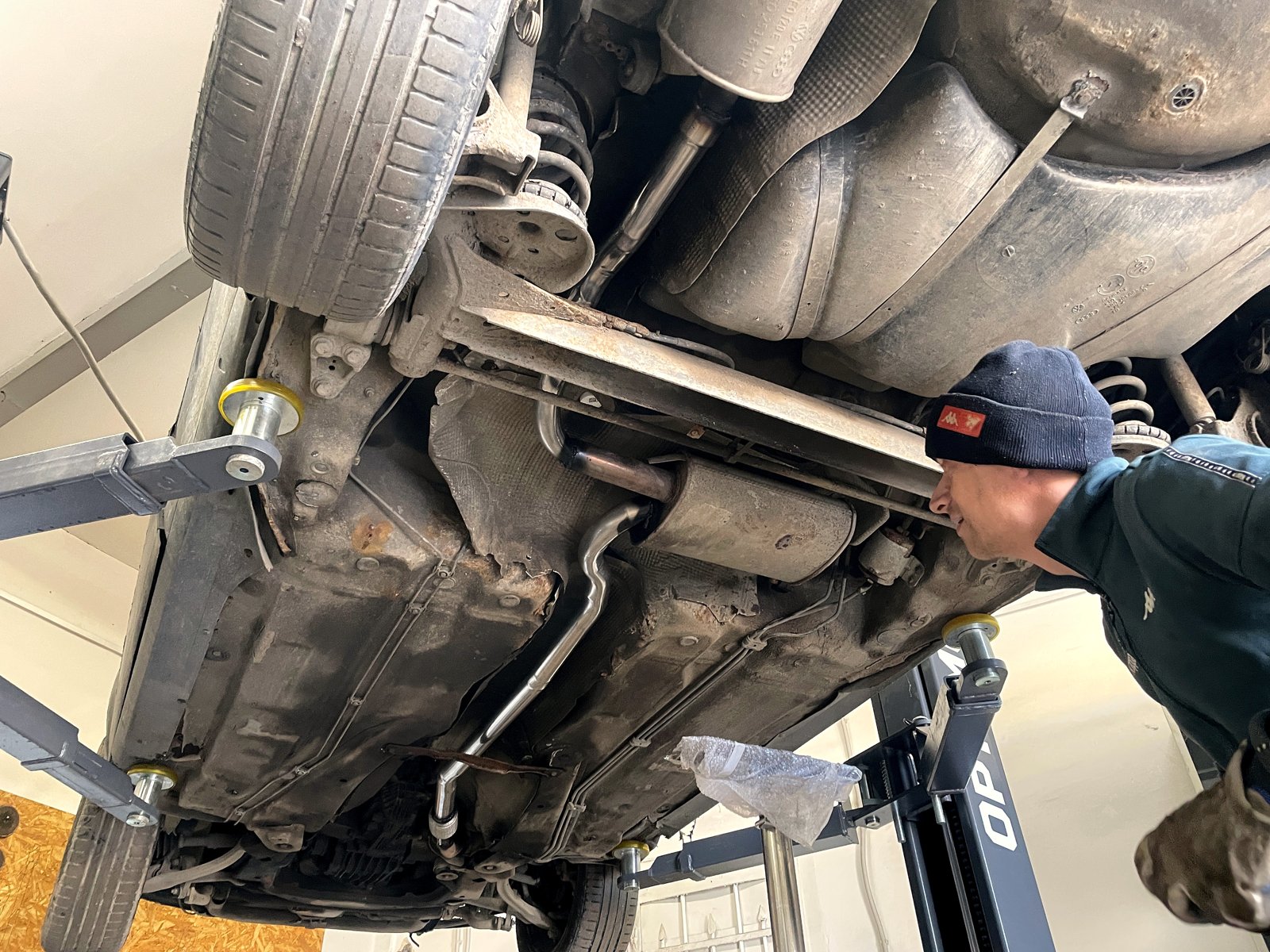 Мастер «Выхлопухи» в шапке осматривает выхлопную трассу VW Passat B5 снизу, вид с кормы