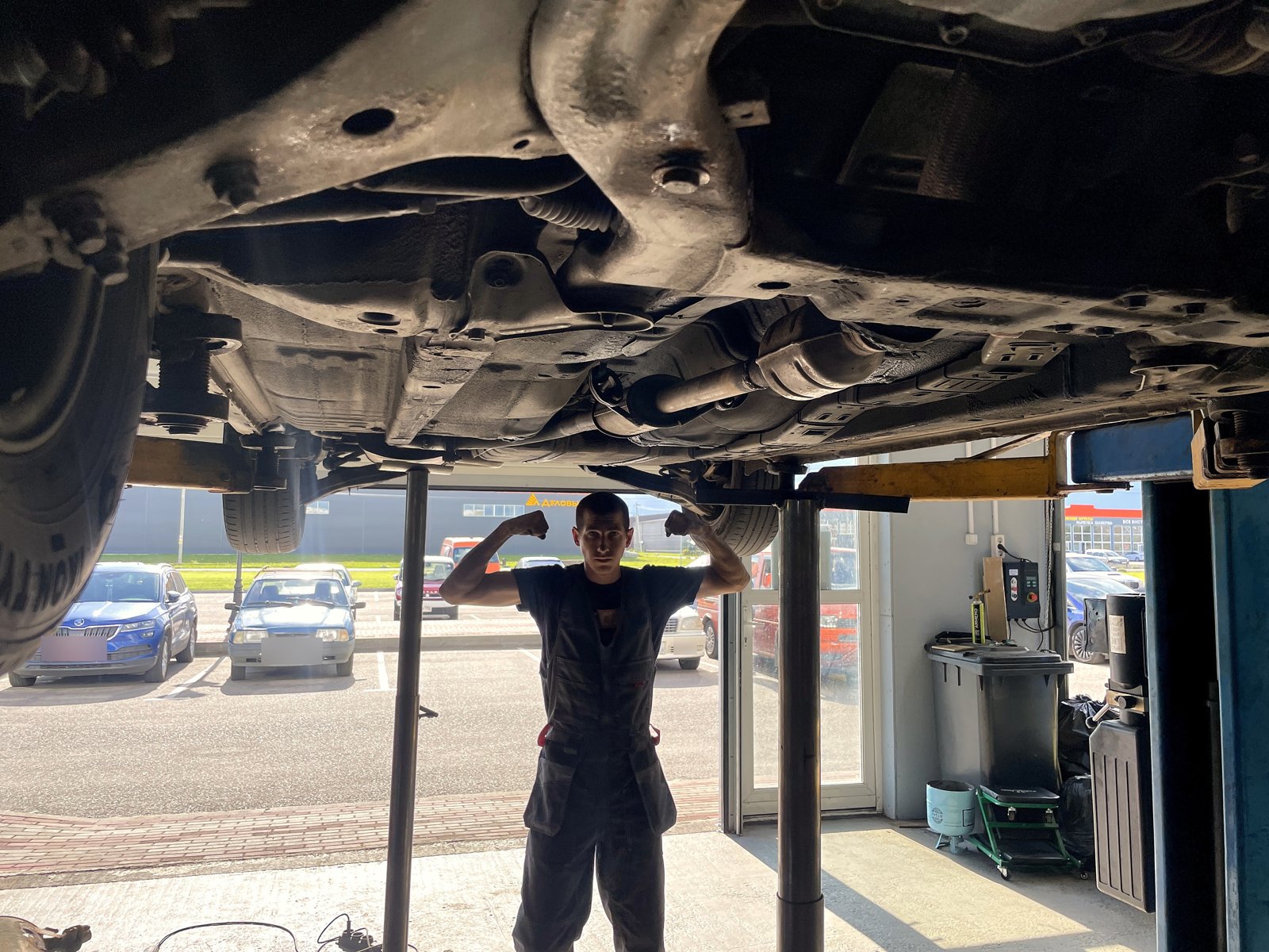 Мастер «Выхлопухи» стоит под поднятым VW Transporter T4, общий вид днища, номера машин на парковке замазаны