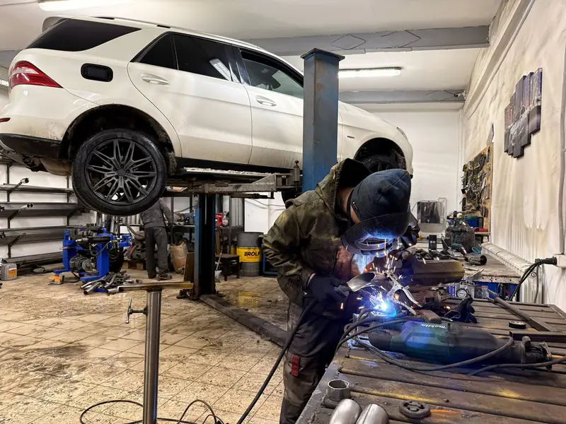 Белый Mercedes ML 350 на подъёмнике в цеху «Выхлопухи», рядом мастер варит на верстаке