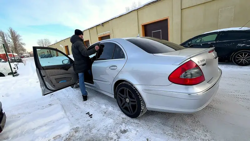 Серебристый Mercedes E-класс W211 на заснеженной площадке у цеха «Выхлопухи», вид сзади сбоку, водитель садится в машину
