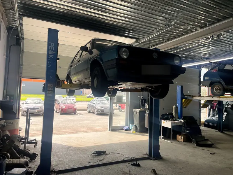 Тёмно-зелёный VW Golf II поднят на двухстоечном подъёмнике в боксе «Выхлопухи» в Тельмана, вид спереди-сбоку