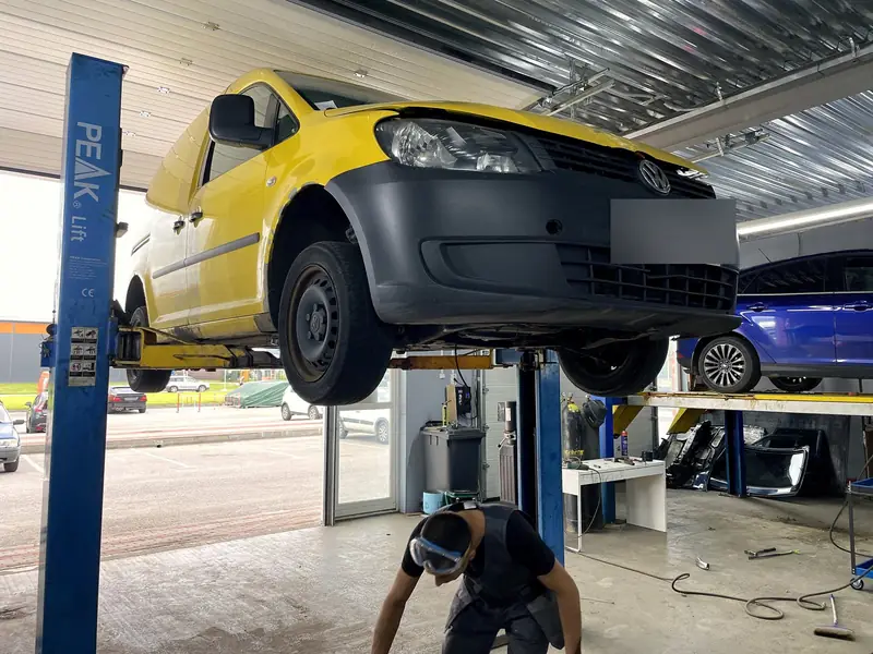 Жёлтый фургон Volkswagen Caddy поднят на двухстоечном подъёмнике в боксе «Выхлопухи», вид спереди снизу