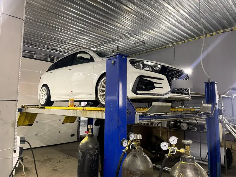 Белая Lada Vesta на подъёмнике в боксе «Выхлопухи» в Тельмана, вид спереди, госномер замазан